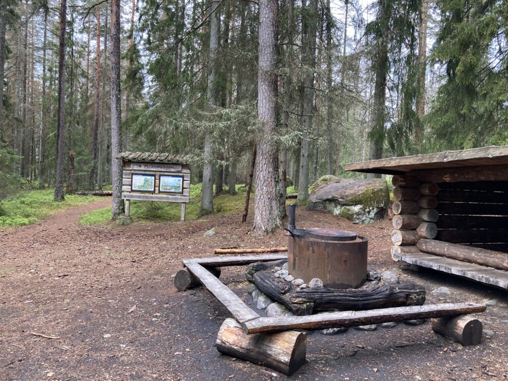 I förgrunden syns en grillplats i skogen med stockar som sittplatser. I högra delen av bilden syns ett vindskydd och i övredelen syns en informationstavla